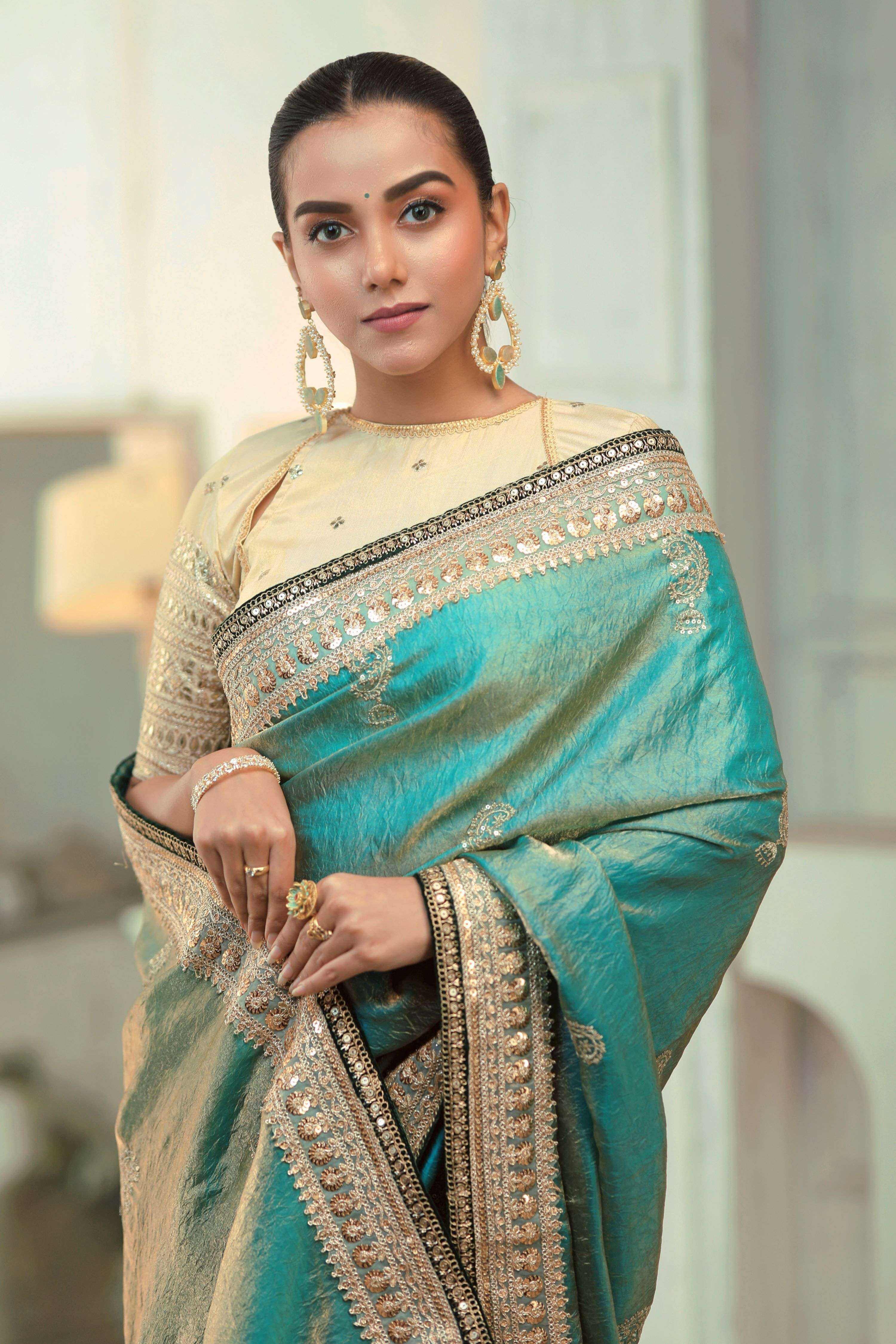 A woman wearing a teal blue saree with champagne blouse, featuring embroidered borders and elegant jewelry.