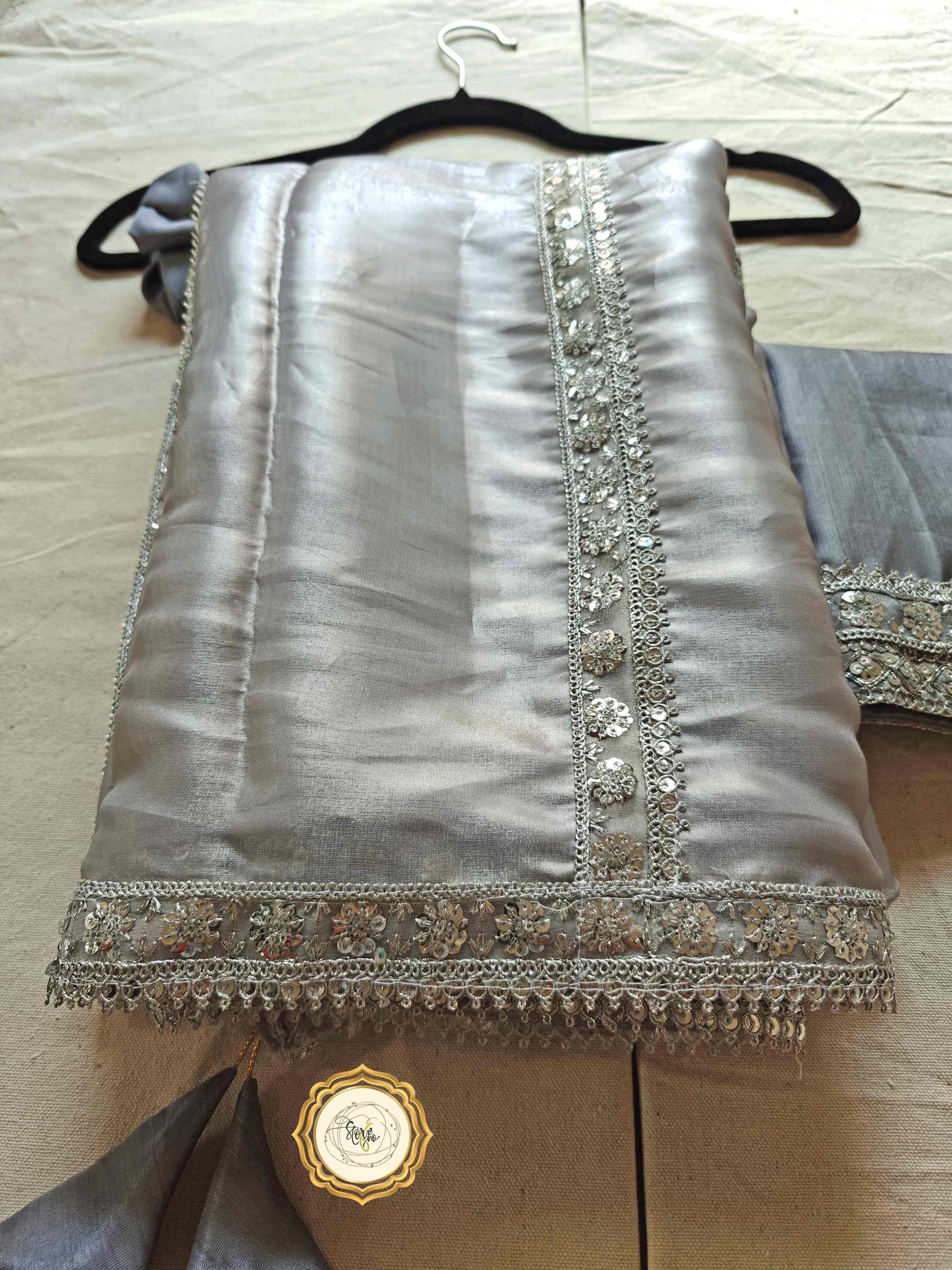 silver organza saree with blouse featuring stunning lace work and cute tassels, displayed neatly on a hanger.