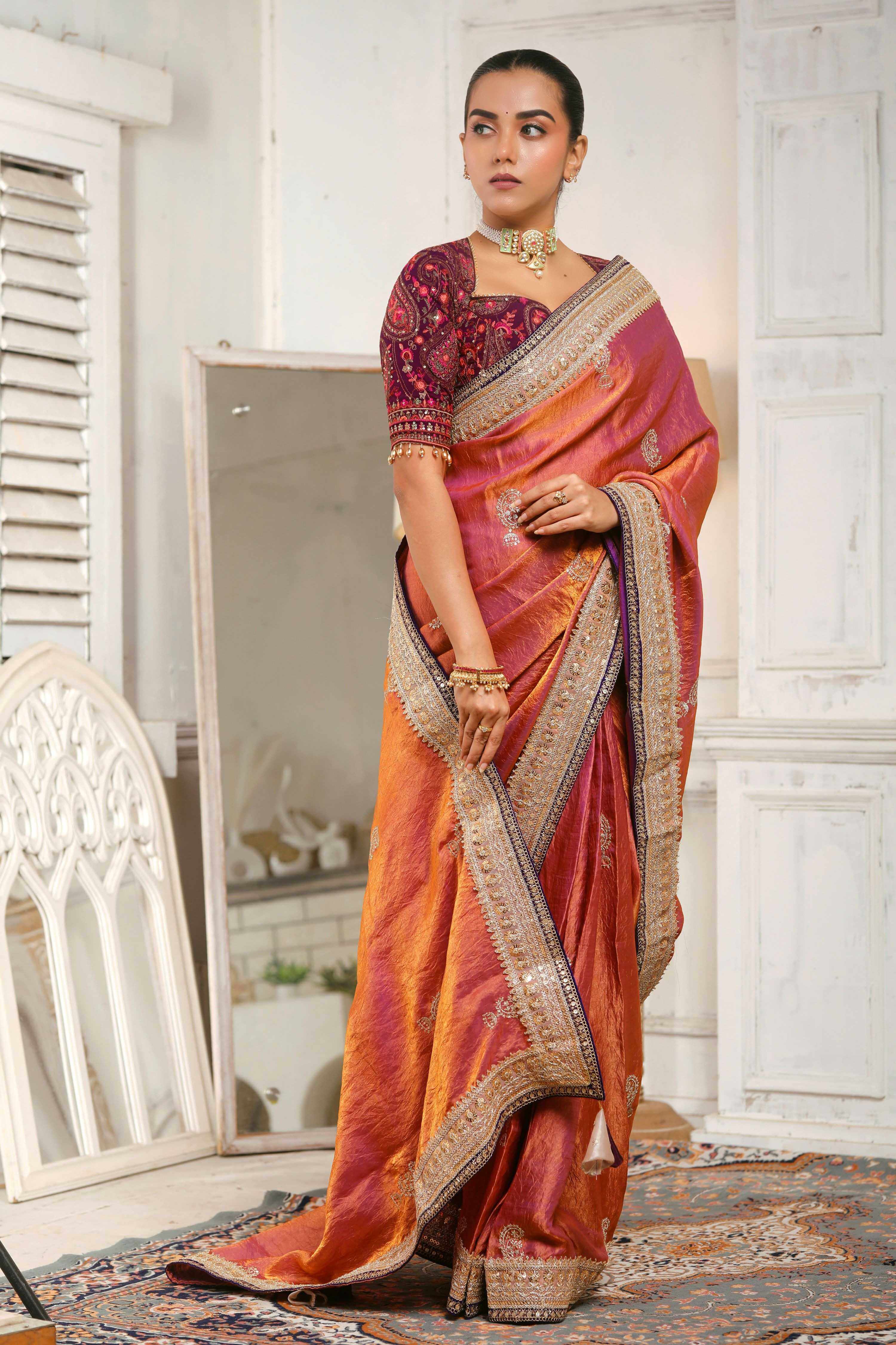 Woman wearing a coral pink crushed tissue saree with embroidered purple and golden borders.